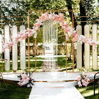 A modern and chic outdoor wedding ceremony setup. The geometric gold frame is adorned with asymmetrical arrangements of pink and white flowers, creating a stylish and contemporary altar on a lush green lawn.