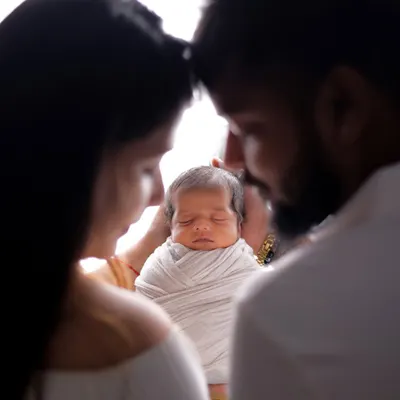 An artistic silhouette shot focusing on the peacefully sleeping newborn. The parents frame their baby, their heads touching in a moment of quiet reverence and love. The backlighting creates a soft, ethereal glow.