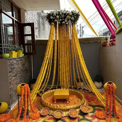 A traditional Haldi setup in a home's courtyard, featuring a canopy of marigold strings, a brass urli for the ceremony, and a floral rangoli on the floor.