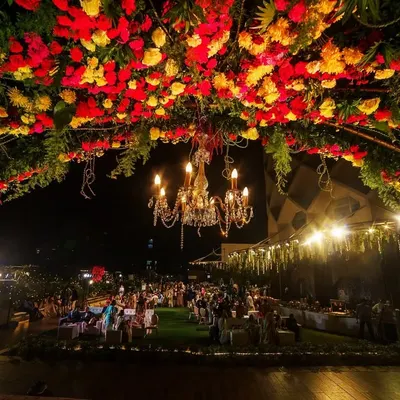 A breathtaking view of a rooftop wedding at night, with a massive floral arch of red and yellow flowers illuminated by a grand chandelier.