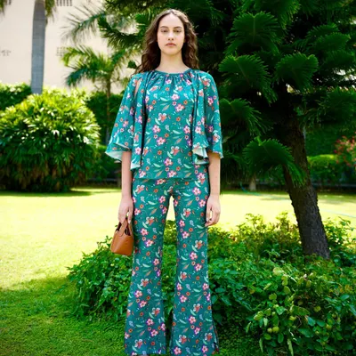 A full-length shot from a summer collection, featuring a green floral co-ord set. The outdoor garden setting and natural light create a fresh, vibrant look perfect for resort wear.