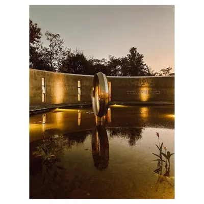 The Dr. Vishnuvardhan Memorial Complex at night, where strategic lighting highlights the texture of the concrete walls and the central ring sculpture. The perfect reflection in the water creates a sense of balance and peace, fulfilling the space's purpose as a place for remembrance. This image also covers media ID 11.