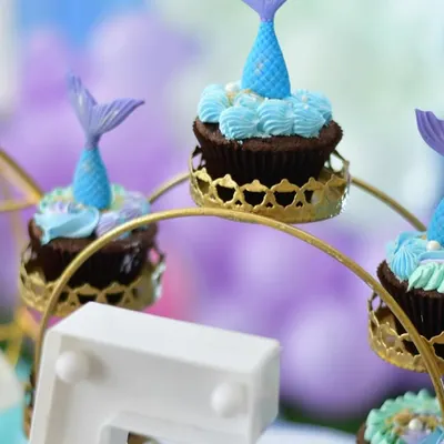 These mermaid-themed cupcakes were part of Saanvi's party. The treats were displayed on a decorative ferris wheel stand, with each cupcake featuring blue and purple frosting and an edible mermaid tail topper.