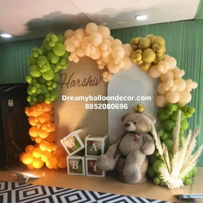 This is a unique jungle-themed decoration I did for a baby celebration. It features a balloon arch in sage green, orange, and beige, with a large teddy bear, B-A-B-Y boxes, and a custom name sign for "Harshiv".