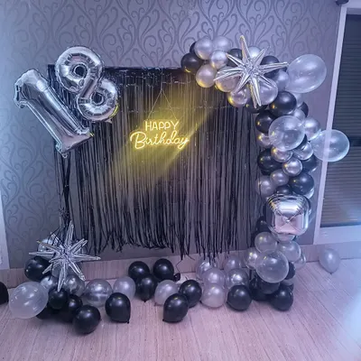 A sleek and modern setup for an 18th birthday. This decoration features a black fringe curtain, a half-arch of black, silver, and clear balloons, starburst foils, and a 'Happy Birthday' neon sign.