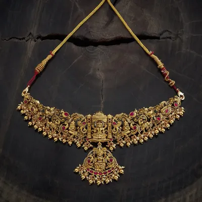 An antique gold-toned temple choker necklace featuring a central pendant of Goddess Lakshmi. The entire piece is detailed with peacock motifs and ruby-colored stones, embodying sacred design and traditional artistry.