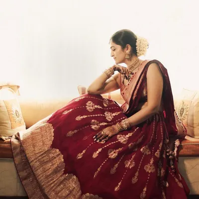A bride, lost in thought, sits by a sunlit window. Her traditional red lehenga spreads around her, creating a beautiful, painterly portrait. This image captures a moment of quiet reflection and timeless elegance.