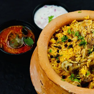 A serving of our wood-fired chicken biryani in a clay pot, accompanied by bowls of tangy Baigan ka Khatta and creamy raita. This is the complete meal experience we deliver for your events.