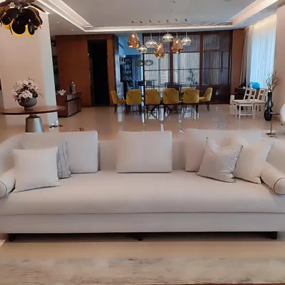 This expansive, timeless white sofa serves as the serene centerpiece of a luxurious open-plan living area. Its clean lines and generous proportions are complemented by an array of textured white cushions, demonstrating our ability to create foundational pieces that exude quiet, sophisticated luxury.
