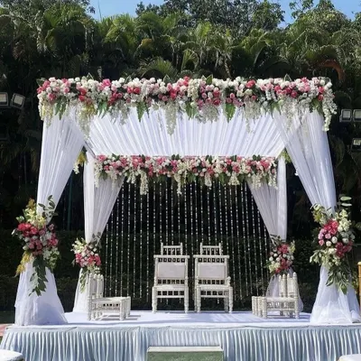 An elegant and classic outdoor wedding mandap design. This setup features a canopy of white drapes, lush pink and white floral arrangements, and delicate crystal hangings, creating a serene and beautiful space for the wedding ceremony.