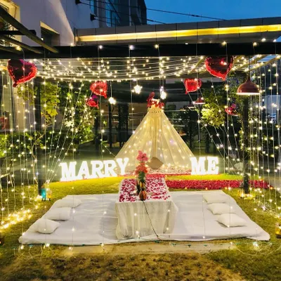 A complete garden proposal package featuring a fairy light cabana for a romantic dinner and a separate teepee tent area. The scene is set with large 'MARRY ME' marquee letters and a path of red rose petals.