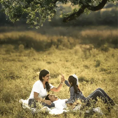 A family enjoys a playful moment during a picnic in a sunlit meadow. The father lies back while the mother and daughter share a high-five, their laughter echoing in the warm, golden light. This is a perfect example of a candid family photoshoot.