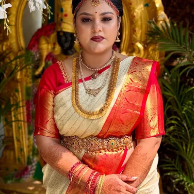 A frontal portrait of this beautiful bride, ready for her muhurtham. The makeup features a classic combination of defined eyes and a deep red lip, designed to harmonize with her traditional cream and red silk saree and gold jewellery.