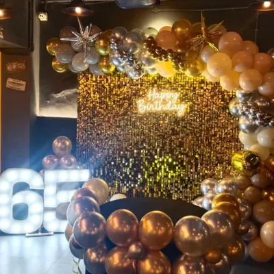 A glamorous setup for a 65th birthday celebration. The decor features a stunning gold sequin wall, a balloon arch in chrome and metallic tones, a 'Happy Birthday' neon sign, and large light up marquee numbers.