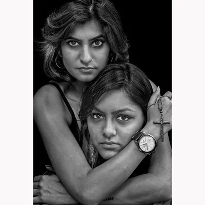 An intense black and white portrait of two women, focusing on their direct gaze and protective embrace. The high-contrast lighting emphasizes the raw emotion and connection between the subjects.