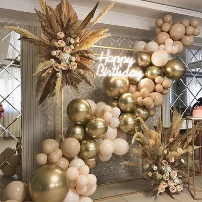 A sophisticated birthday backdrop featuring a gold frame against a mirrored wall. The balloon garland in nude, gold, and blush tones is paired with dried pampas grass and a "Happy Birthday" neon sign for a trendy, bohemian-luxe feel.
