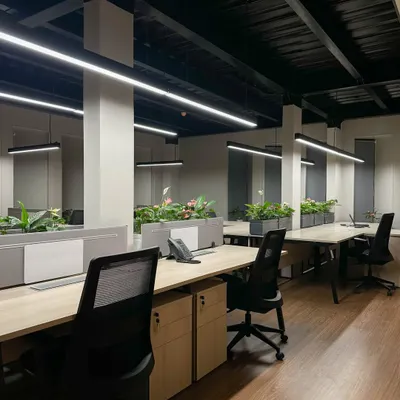 A view across the open-plan work floor, featuring rows of workstations with integrated planters. The industrial-inspired exposed ceiling is fitted with modern linear lighting to ensure a bright and energizing environment.