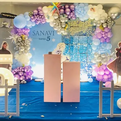 A magical Frozen theme for a 5th birthday. The backdrop features a shimmering blue waterfall effect, surrounded by a balloon arch in shades of purple, blue, and silver, with cutouts of Anna and Elsa.
