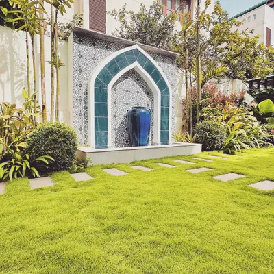 A magnificent Moroccan-style archway, tiled in deep blue, frames a large urn, creating a dramatic and serene water feature. Set against a backdrop of patterned tiles and surrounded by a lush lawn, it serves as an elegant garden centerpiece.