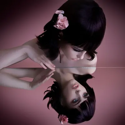 A creative beauty portrait showing a model with a sleek black bob and a pink flower in her hair, gazing at her own reflection in a mirror. This image showcases my technical skill with lighting and composition for artistic and conceptual photoshoots.