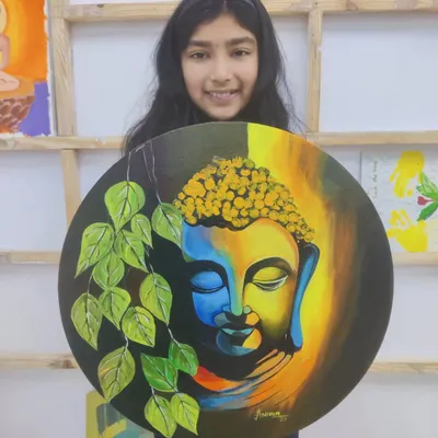 A young student proudly holds her beautifully finished acrylic painting of Buddha on a round canvas, a piece that radiates peace and serenity.