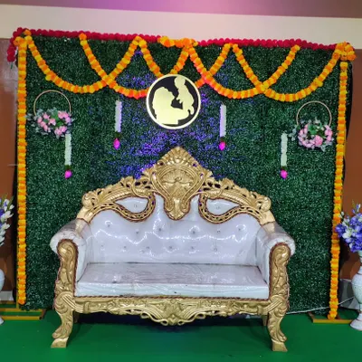 This is a traditional decoration I provide for events like baby showers. It features a green wall backdrop, marigold flower garlands, and an ornate white and gold sofa for seating.