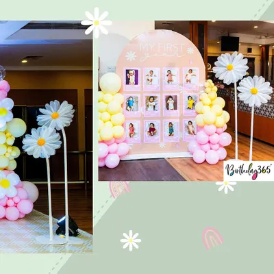 A sweet and cheerful 'My First Year' photo board with a daisy theme. The arched display is decorated with pastel pink and yellow balloons and features large, charming daisy props on either side.