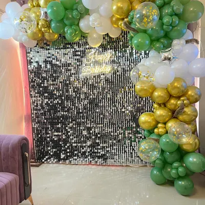 A stylish birthday backdrop featuring a shimmering silver sequin wall. It is framed by a green, gold, and white balloon arch and includes a 'Happy Birthday' neon sign.