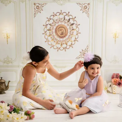 A sweet, candid moment between two sisters. The older sister gently adjusts the younger one's hair bow in our elegant, light-filled studio, showing a quiet moment of care.
