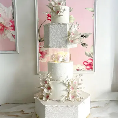 A grand creation for life’s grandest moments. This five-tiered white anniversary cake is a showstopper, featuring hexagonal and square tiers, intricate textures, and delicate, handcrafted sugar flowers.