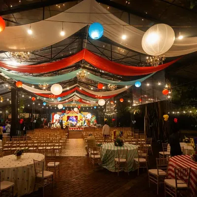 A wide shot of a spectacular carnival-themed event at night. The venue is transformed with colorful draped fabrics, paper lanterns, and string lights, creating the festive atmosphere of a big top tent.