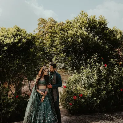 A beautiful wide shot of a couple in coordinated dark green traditional outfits, standing in a lush garden. The natural lighting and rich colors make this a stunning pre-wedding portrait.