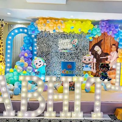 A vibrant Cocomelon-themed first birthday party. This stage setup features a silver shimmer wall, character cutouts, a tall giraffe prop, and large marquee letters.