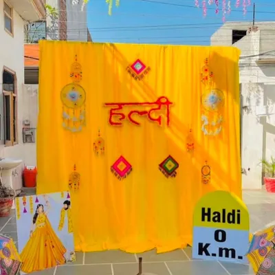 A classic and fun outdoor Haldi ceremony setup. The backdrop is made of bright yellow drapes with a 'Haldi' sign in Hindi, accented with traditional hangings and quirky props like the 'Haldi 0 Km' milestone.