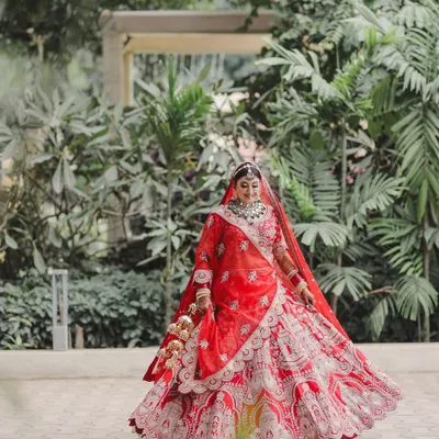 A full-length shot of the bride in her magnificent red lehenga, capturing the grandeur of her attire against a lush green backdrop. The twirl of the skirt shows the intricate detail and craftsmanship.