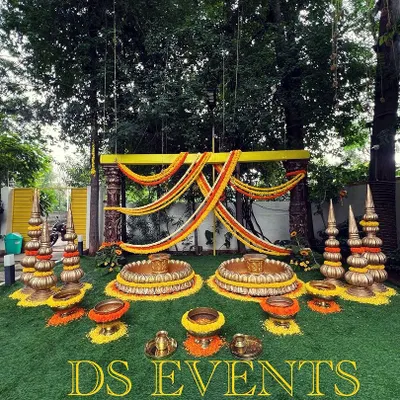 An elaborate outdoor setup for a Haldi or Pellikuthuru ceremony. This design features two large lotus-shaped urlis for seating, surrounded by traditional brass decor elements and marigold flower arrangements.