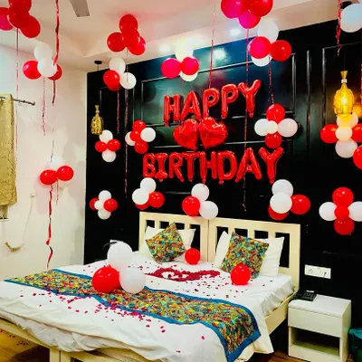 A classic romantic birthday bedroom decoration. The room is filled with red and white balloons, a large "Happy Birthday" foil balloon set above the bed, and rose petals scattered for that lovely touch.