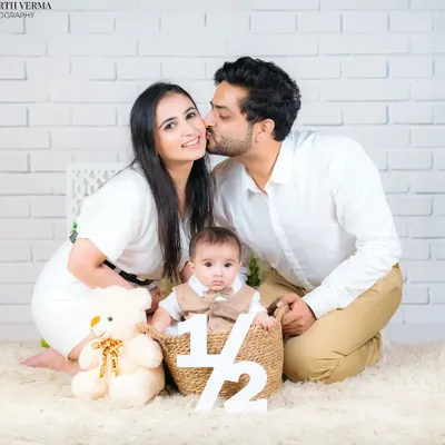 A sweet half-birthday celebration photo. The father's kiss on the mother's cheek while they surround their baby creates a moment of pure family love.