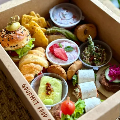 This is an example of my curated Gourmet Food Boxes. It's a personal feast, featuring a mini slider, a savory croissant, truffle edamame pockets, fresh dips, and other artisanal bites, all packed into one convenient box.
