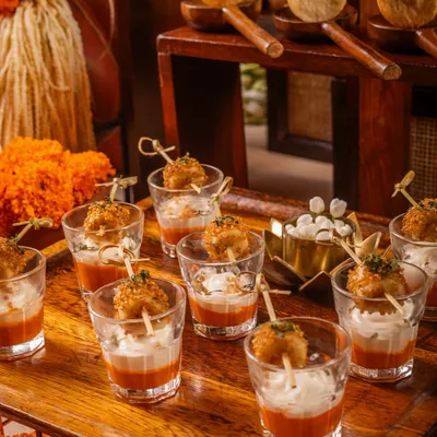 An array of our signature appetizers, perfect for a festive celebration. This tray features crispy bites in shot glasses with a layered dip and pani puris on custom wooden spoons, all set against a backdrop of vibrant marigold flowers.