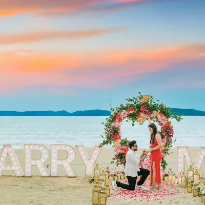 A picture-perfect beach proposal captured during a pre-wedding shoot. The "MARRY ME" marquee lights, floral arch, and stunning sunset sky create an unforgettable romantic moment.