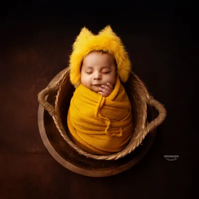 A classic and adorable posed newborn photo. The baby is comfortably swaddled in a vibrant yellow wrap and a cute, furry hat, nestled safely in a woven basket for a perfect, sleepy portrait.