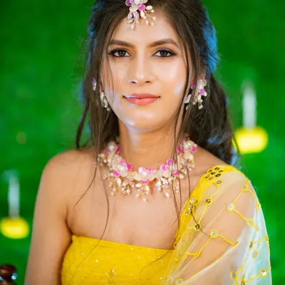 The Haldi glow is real. This is a beautiful close-up portrait of a bride-to-be, adorned with delicate floral jewelry, looking directly at the camera with a serene smile. The vibrant green background makes her yellow outfit pop.