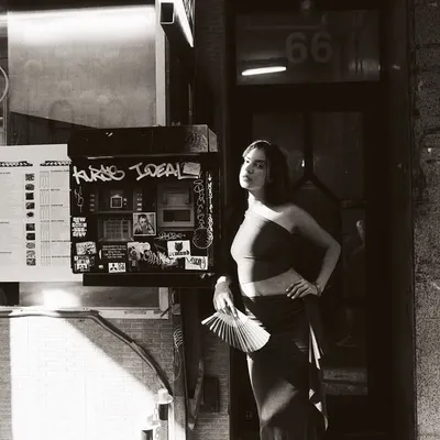 A black and white street portrait. The harsh sunlight creates a dramatic diagonal shadow, cutting through the frame. Monochrome enhances the textures of the street and the play of light, making it feel like a still from a classic film.
