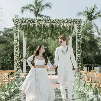 A fairytale wedding in a pristine all-white theme. This shot captures the couple walking down a mirrored aisle, surrounded by lush greenery and palm trees.
