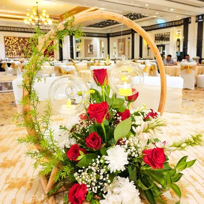 A unique and rustic centerpiece featuring a wooden hoop entwined with greenery. The arrangement of red and white flowers is accented with hanging glass orbs containing small candles, adding a touch of warmth and charm.