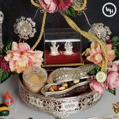 This opulent silver-plated tray is a centerpiece gift for Diwali. It features silver Lakshmi-Ganesh idols for blessings, a crystal jar for sweets, and a peanut-shaped container for dry fruits, all adorned with lush pink flowers.