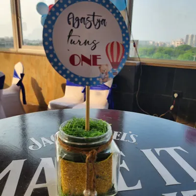 A charming table centerpiece for a hot air balloon themed first birthday. The personalized "Agastya turns ONE" sign is placed in a jar filled with glitter and moss, adding a touch of rustic charm.