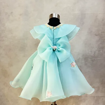 A custom dress in a beautiful shade of aqua organza. The design features a Peter Pan collar, delicate floral embroidery, and a large, statement bow at the back.