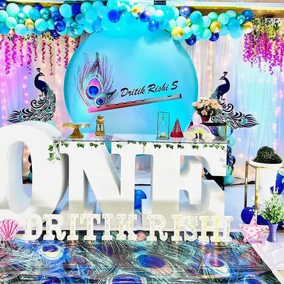 This is a complete custom setup I did for a first birthday photoshoot with a Krishna theme. I designed and arranged everything you see, including the large 'ONE' table, the peacock feather printed backdrop and floor mat, and the blue and purple balloon arch.